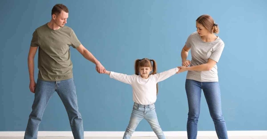 A man and woman each holding an arm of their young daughter as if they are pulling her in opposite directions