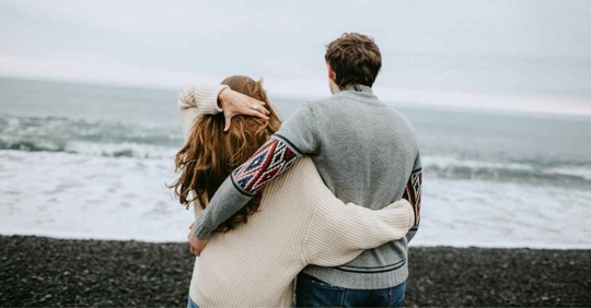 A man and woman standing at the beach looking at the ocean with their arms around each other