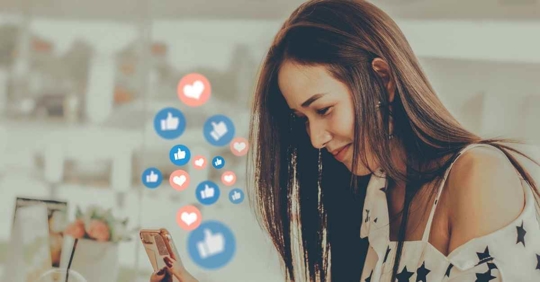 A woman looking at her phone with various social icons floating around her. The blue thumb "like" button and red heart "love" button appear.
