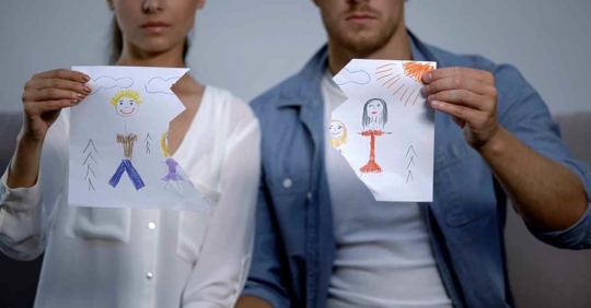 A man and woman sitting next to each other. Each holds a ripped piece of paper with a drawing of a child on it