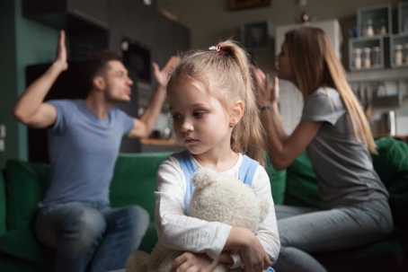 Young girl holding a teddy bear, parents fighting behind