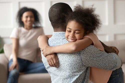 Young girl hugging a man while a woman sitting on a couch smiles in the background.